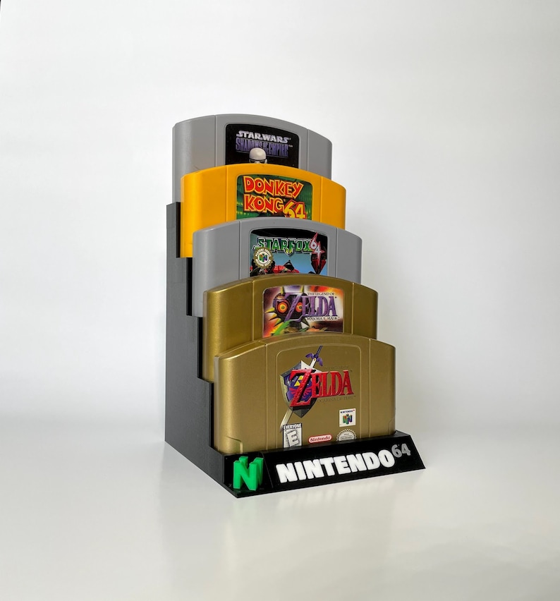 Puede incluir: Un soporte negro para cartuchos de juegos de Nintendo 64 con cuatro cartuchos de juegos. Los cartuchos son, de arriba a abajo: Star Wars Shadows of the Empire, Donkey Kong 64, Starfox 64 y The Legend of Zelda: Ocarina of Time.