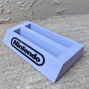 May include: A light purple plastic Nintendo game cartridge holder with two slots. The holder has the word "Nintendo" printed in black on the front.