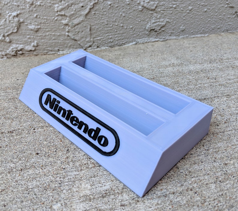 May include: A light purple plastic Nintendo game cartridge holder with two slots. The holder has the word "Nintendo" printed in black on the front.