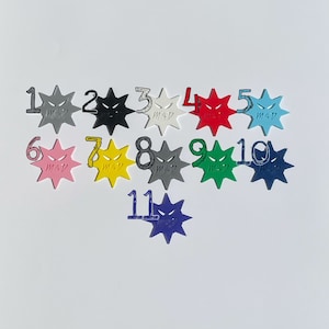 Puede incluir: Once fichas de colores en forma de estrella con un dise&ntilde;o de cara enojada en blanco y negro. Cada ficha est&aacute; numerada del 1 al 11.