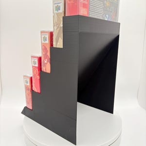 May include: A black, stepped display stand holding several Nintendo 64 game boxes. The boxes are red and beige, with game titles and logos visible. The stand is on a white, rotating platform.