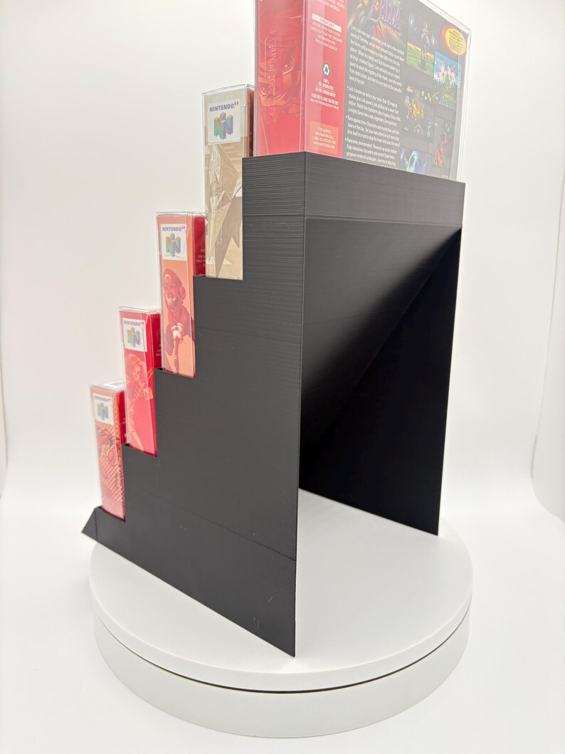 May include: A black, stepped display stand holding several Nintendo 64 game boxes. The boxes are red and beige, with game titles and logos visible. The stand is on a white, rotating platform.
