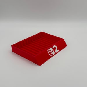 Pu&ograve; includere: Un vassoio rosso rettangolare con una serie di scanalature parallele. Il vassoio ha un design inclinato e presenta il testo bianco "Switch 2" sul lato. Il prodotto &egrave; probabilmente progettato per organizzare o esporre oggetti.