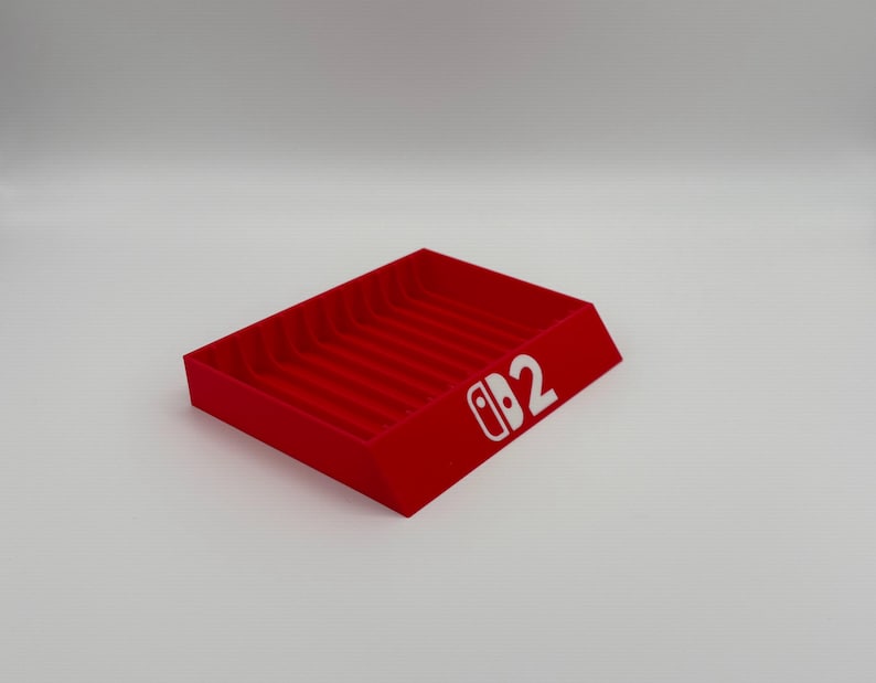 Pu&ograve; includere: Un vassoio rosso rettangolare con una serie di scanalature parallele. Il vassoio ha un design inclinato e presenta il testo bianco "Switch 2" sul lato. Il prodotto &egrave; probabilmente progettato per organizzare o esporre oggetti.