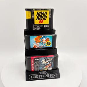 K&ouml;nnte beinhalten: Ein schwarzer Sega Genesis-Spielkassetten-Displayst&auml;nder mit drei Spielkassetten. Die oberste Kassette ist "Road Rash II", die mittlere ist "Sonic the Hedgehog" und die untere ist "Batman Forever". Das Sega Genesis-Logo ist sichtbar.
