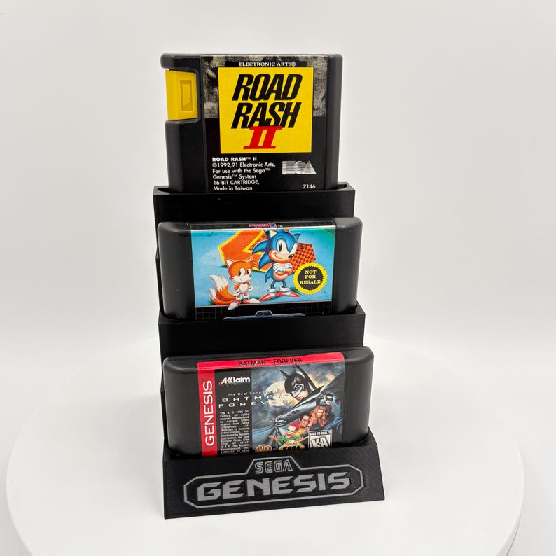 K&ouml;nnte beinhalten: Ein schwarzer Sega Genesis-Spielkassetten-Displayst&auml;nder mit drei Spielkassetten. Die oberste Kassette ist "Road Rash II", die mittlere ist "Sonic the Hedgehog" und die untere ist "Batman Forever". Das Sega Genesis-Logo ist sichtbar.