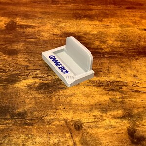 May include: White plastic stand with the words "GAMEBOY" printed on the front. The stand is designed to hold a Game Boy handheld gaming device.