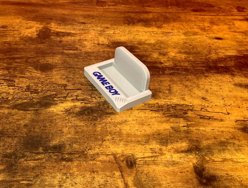 May include: White plastic stand with the words "GAMEBOY" printed on the front. The stand is designed to hold a Game Boy handheld gaming device.