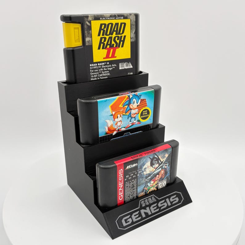 K&ouml;nnte beinhalten: Ein schwarzer Display-St&auml;nder mit drei Sega Genesis Videospiel-Cartridges. Die oberste Cartridge ist "Road Rash II", die mittlere zeigt Sonic the Hedgehog und die untere "Batman Forever". Der St&auml;nder hat das Sega Genesis Logo.