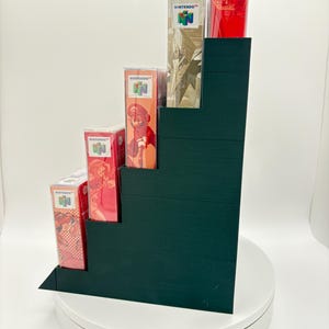 May include: A dark green stepped display stand holding several Nintendo 64 game boxes and a red controller. The boxes feature colorful game art and the Nintendo logo. The stand is on a white rotating platform.