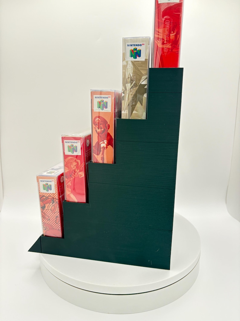 May include: A dark green stepped display stand holding several Nintendo 64 game boxes and a red controller. The boxes feature colorful game art and the Nintendo logo. The stand is on a white rotating platform.