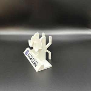 May include: A white 3D-printed Nintendo GameCube logo and character display. The display features the GameCube logo and a stylized character design. The base of the display has the words "NINTENDO GAME CUBE" printed on it.