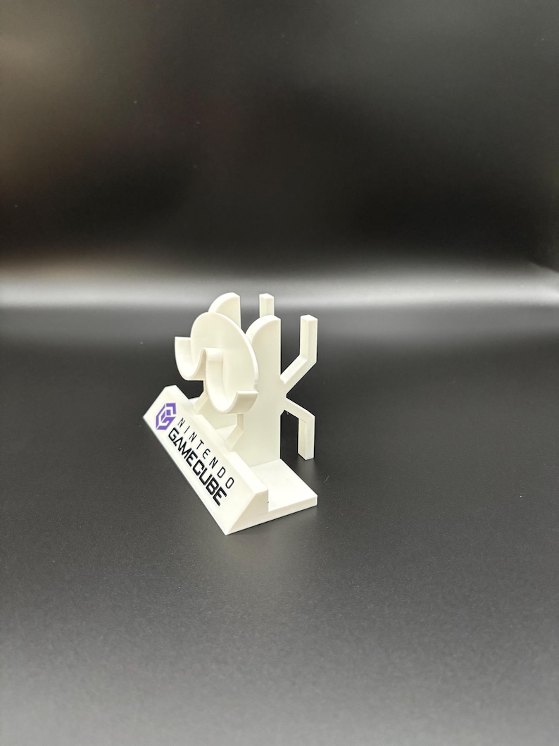 May include: A white 3D-printed Nintendo GameCube logo and character display. The display features the GameCube logo and a stylized character design. The base of the display has the words "NINTENDO GAME CUBE" printed on it.