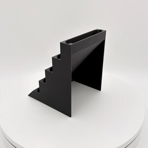 K&ouml;nnte beinhalten: Ein schwarzer, geometrischer Pr&auml;sentationsst&auml;nder mit Stufen. Der St&auml;nder hat eine Reihe von aufsteigenden Stufen auf einer Seite und eine rechteckige Rinne oben. Das Design ist minimalistisch und modern, mit einer einfarbigen schwarzen Farbe.