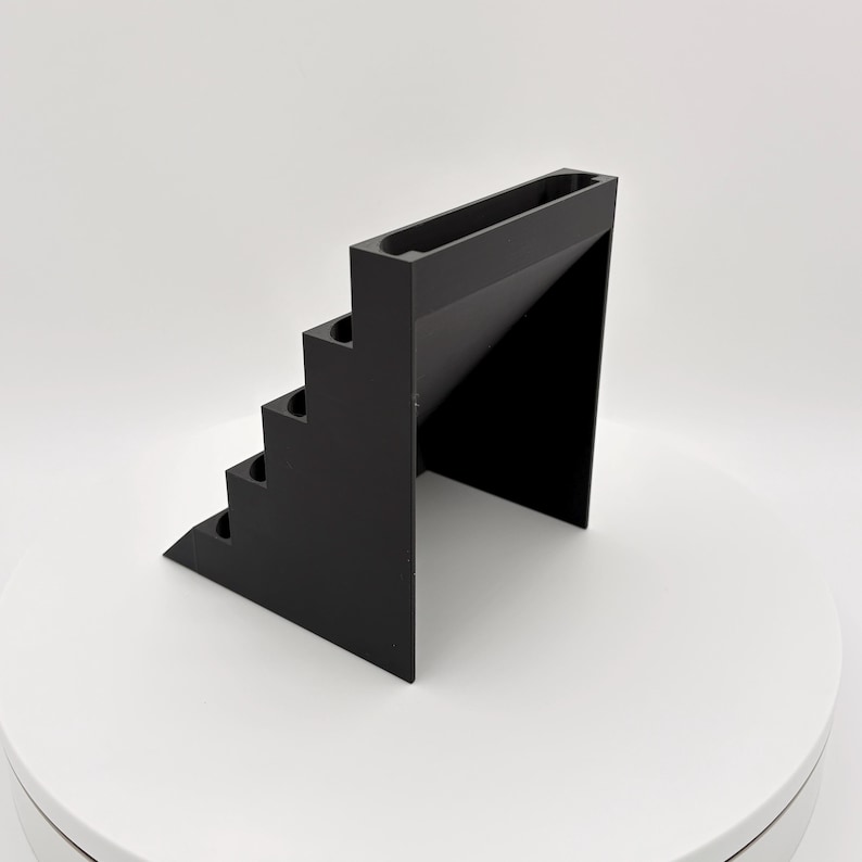 K&ouml;nnte beinhalten: Ein schwarzer, geometrischer Pr&auml;sentationsst&auml;nder mit Stufen. Der St&auml;nder hat eine Reihe von aufsteigenden Stufen auf einer Seite und eine rechteckige Rinne oben. Das Design ist minimalistisch und modern, mit einer einfarbigen schwarzen Farbe.