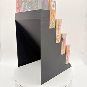 May include: A black, stepped display stand holding several video game cartridges and boxes. The stand is made of a solid material and has a modern, geometric design. The games are visible on the steps.