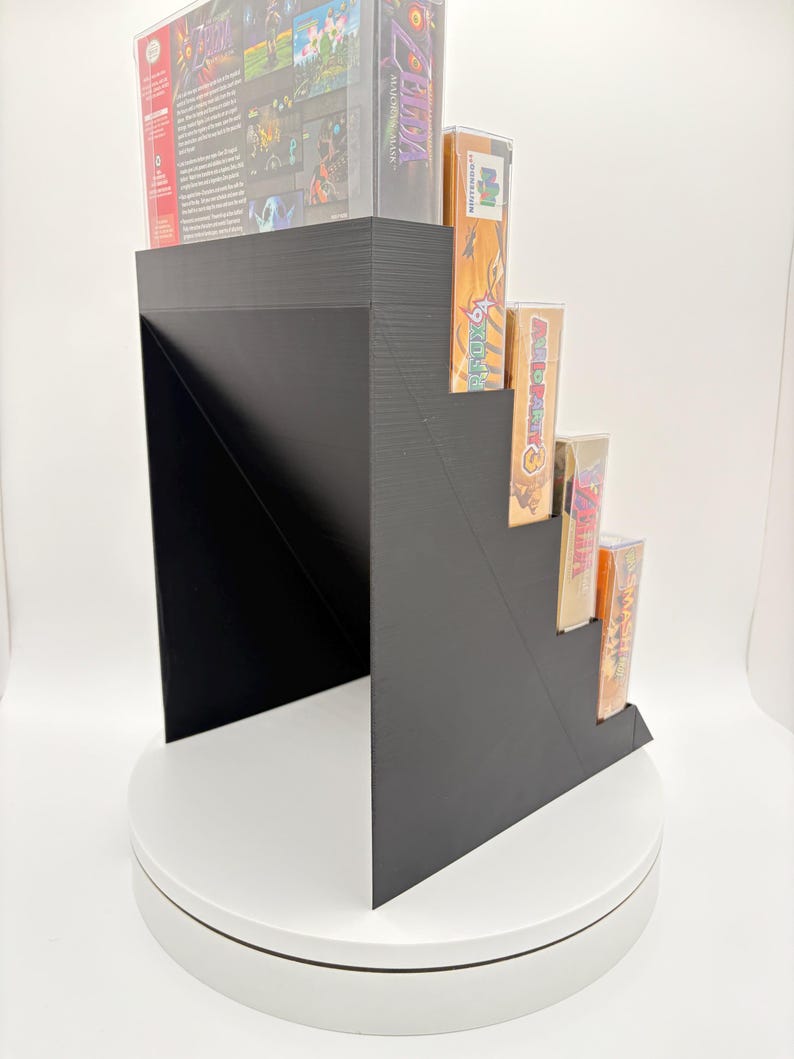 May include: A black, stepped display stand holding several video game cartridges and boxes. The stand is made of a solid material and has a modern, geometric design. The games are visible on the steps.