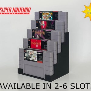 May include: A black Super Nintendo game cartridge holder with five slots, displaying various game cartridges, including Super Mario All-Stars, The Secret of Mana, Final Fantasy III, Final Fantasy IV, and Chrono Trigger. The holder is available in sizes from 2 to 6 slots. "AVAILABLE IN 2-6 SLOTS" is printed below the holder.