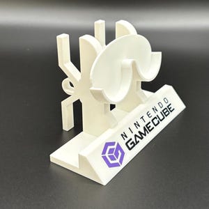 May include: A white 3D-printed Nintendo GameCube stand. The stand features the GameCube logo in purple and the words "NINTENDO GAMECUBE" in black. The stand has a unique design with curved and angular elements.