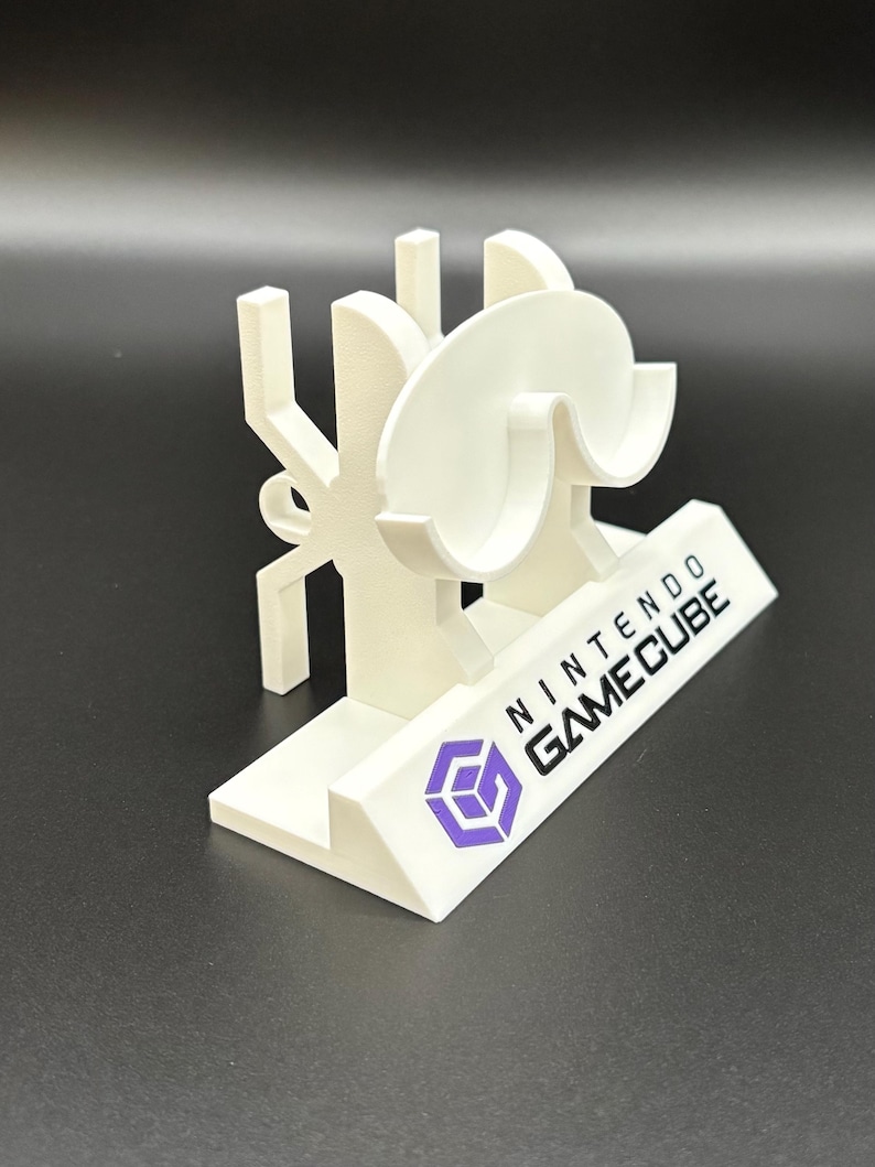 May include: A white 3D-printed Nintendo GameCube stand. The stand features the GameCube logo in purple and the words "NINTENDO GAMECUBE" in black. The stand has a unique design with curved and angular elements.