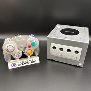 May include: A silver Nintendo GameCube console and matching controller. The controller has a gray body with colored buttons and a joystick. The console has a disc tray and several ports. The controller rests on a white stand with the Nintendo GameCube logo.