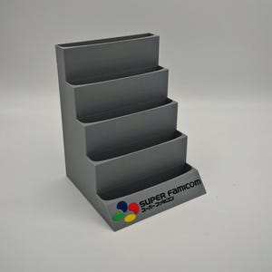 Puede incluir: Un organizador gris escalonado con cuatro estantes. La base presenta el logotipo "SUPER FAMICOM" en ingl&eacute;s y japon&eacute;s, con un emblema colorido. El organizador est&aacute; dise&ntilde;ado para guardar art&iacute;culos y tiene un dise&ntilde;o escalonado.