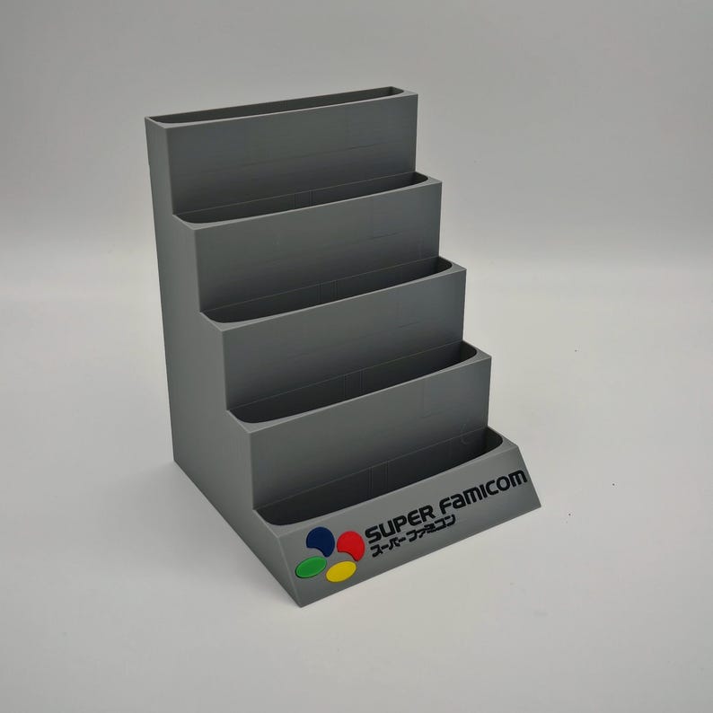 Puede incluir: Un organizador gris escalonado con cuatro estantes. La base presenta el logotipo "SUPER FAMICOM" en ingl&eacute;s y japon&eacute;s, con un emblema colorido. El organizador est&aacute; dise&ntilde;ado para guardar art&iacute;culos y tiene un dise&ntilde;o escalonado.