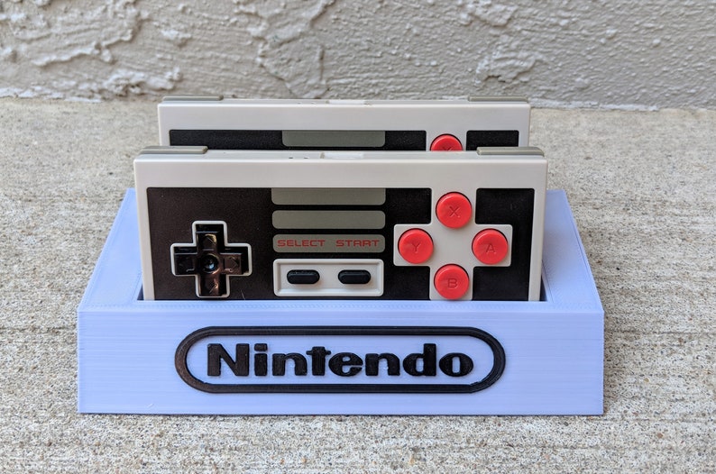 May include: A light blue plastic stand with the word "Nintendo" printed on the front. Two gray and black Nintendo Entertainment System controllers are sitting in the stand.