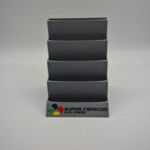 Puede incluir: Un organizador gris en niveles para cartuchos de videojuegos. La base presenta el logotipo de Super Famicom en color, con el texto "SUPER FAMICOM" y caracteres japoneses. El organizador tiene cuatro compartimentos abiertos.