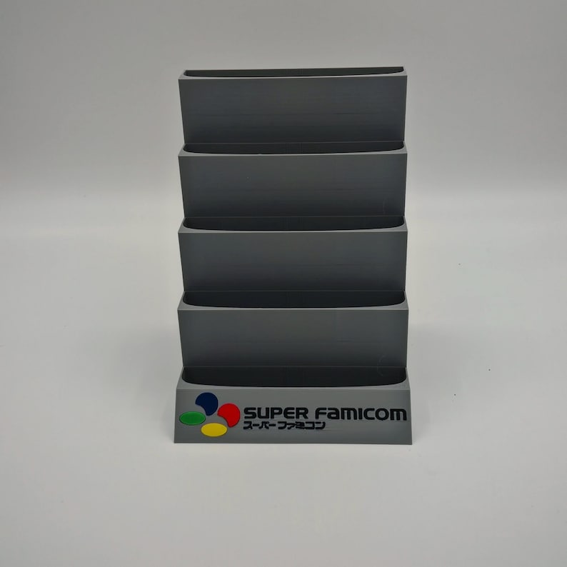 Puede incluir: Un organizador gris en niveles para cartuchos de videojuegos. La base presenta el logotipo de Super Famicom en color, con el texto "SUPER FAMICOM" y caracteres japoneses. El organizador tiene cuatro compartimentos abiertos.