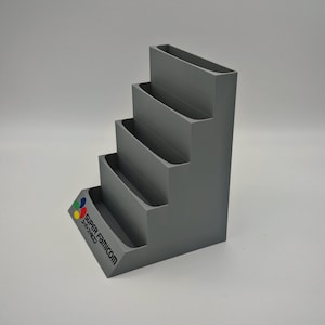 Puede incluir: Un expositor gris escalonado con cuatro niveles y una base inclinada. La base presenta el logotipo y el texto de Super Famicom en varios colores. El soporte est&aacute; dise&ntilde;ado para sujetar y exhibir art&iacute;culos.