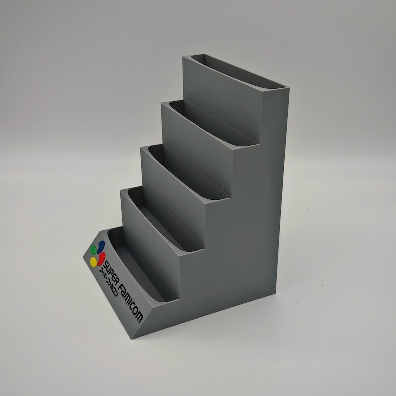 Puede incluir: Un expositor gris escalonado con cuatro niveles y una base inclinada. La base presenta el logotipo y el texto de Super Famicom en varios colores. El soporte est&aacute; dise&ntilde;ado para sujetar y exhibir art&iacute;culos.