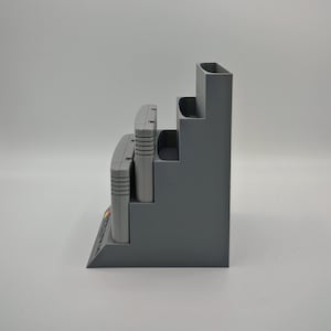 Puede incluir: Un organizador gris escalonado dise&ntilde;ado para guardar cartuchos de videojuegos. El organizador tiene tres niveles, cada uno dimensionado para contener un cartucho. La base del organizador tiene un peque&ntilde;o estante en &aacute;ngulo. Los cartuchos son grises y tienen etiquetas coloridas.