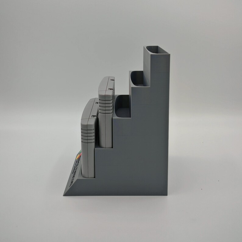 Puede incluir: Un organizador gris escalonado dise&ntilde;ado para guardar cartuchos de videojuegos. El organizador tiene tres niveles, cada uno dimensionado para contener un cartucho. La base del organizador tiene un peque&ntilde;o estante en &aacute;ngulo. Los cartuchos son grises y tienen etiquetas coloridas.