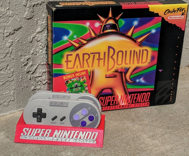 May include: A red and white Super Nintendo Entertainment System game cartridge case with a colorful image of a robot character on the front. The case is labeled "EarthBound" and "Only For Nintendo". Two gray Super Nintendo controllers are stacked on top of a red Super Nintendo stand.