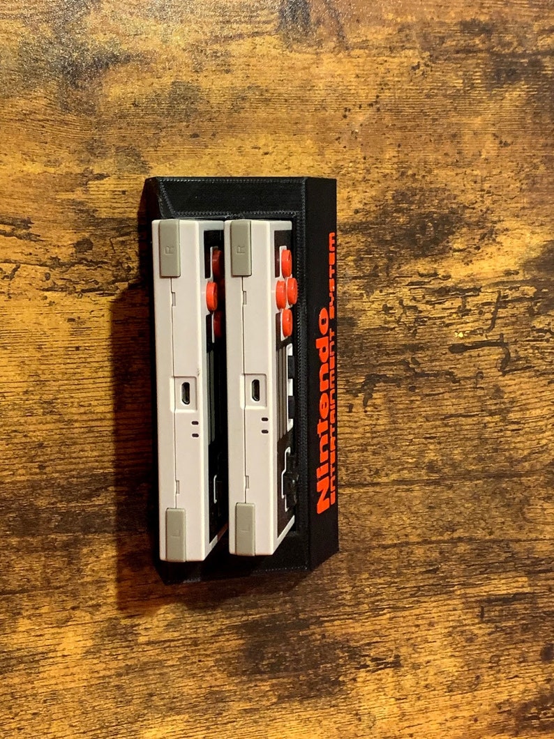 May include: Two white Nintendo Entertainment System controllers are mounted in a black wall mount. The controllers have red buttons and a black and white design.