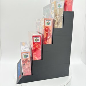 May include: A black, stepped display stand holding several Nintendo 64 game boxes. The boxes feature colorful game art and the Nintendo logo. The stand is on a white surface, showcasing the games.