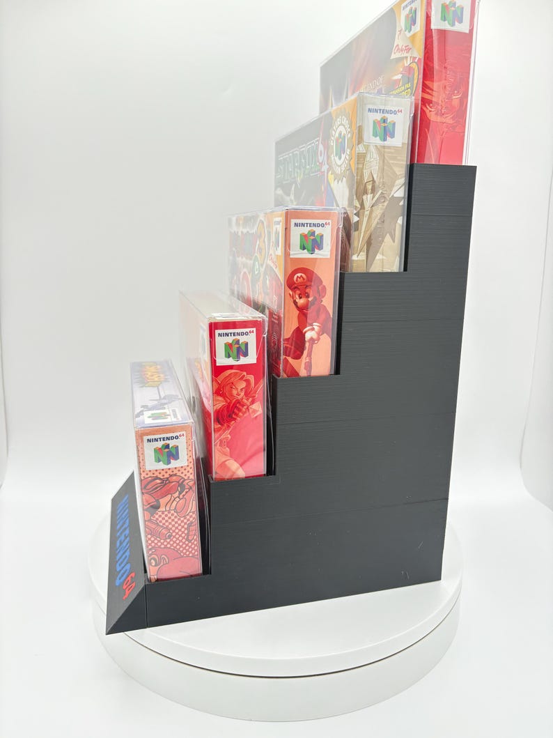May include: A black, stepped display stand holding several Nintendo 64 game boxes. The boxes feature colorful game art and the Nintendo logo. The stand is on a white surface, showcasing the games.