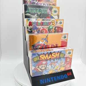 May include: A black display stand holding five Nintendo 64 video game boxes. The games include "Super Smash Bros," "The Legend of Zelda," "Mario Party 3," and "Star Fox 64." The stand has the Nintendo 64 logo in blue.