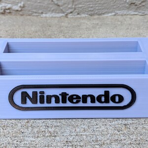 May include: A light purple plastic game cartridge holder with two slots and the Nintendo logo on the front.