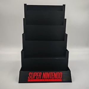 May include: A black cardboard display stand with multiple shelves. The stand has the words "SUPER NINTENDO ENTERTAINMENT SYSTEM" printed in red on the front. The stand is designed to hold items for display.