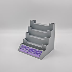 Puede incluir: Un soporte de pl&aacute;stico gris con cinco estantes para guardar cartuchos de juegos de Super Nintendo. El soporte tiene las palabras "SUPER NINTENDO ENTERTAINMENT SYSTEM" impresas en la parte delantera.