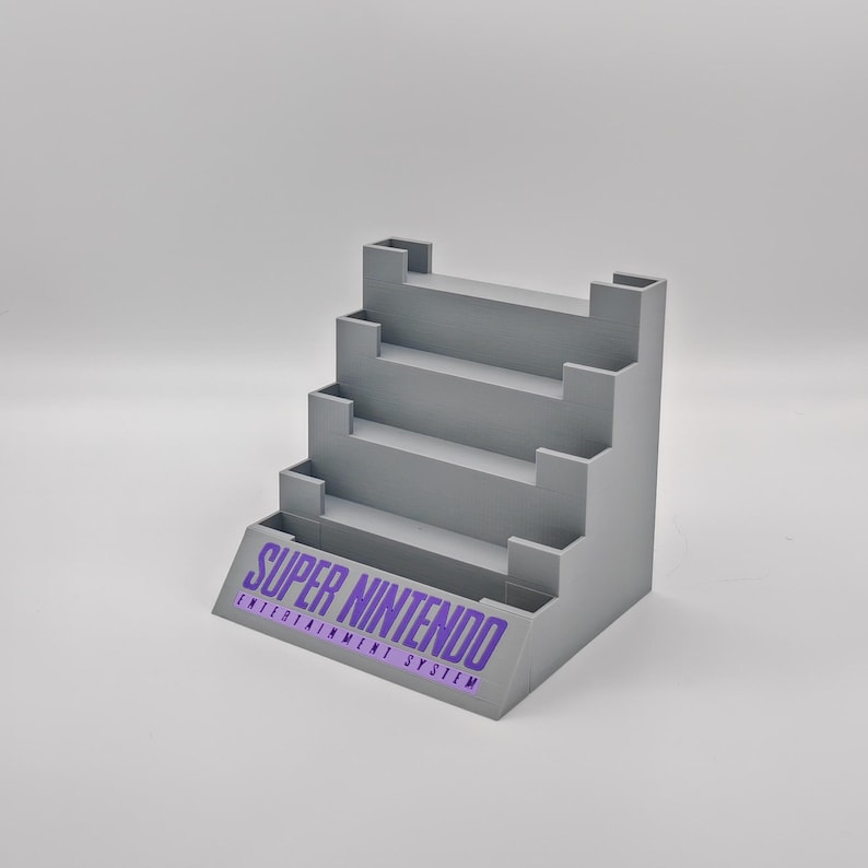 Puede incluir: Un soporte de pl&aacute;stico gris con cinco estantes para guardar cartuchos de juegos de Super Nintendo. El soporte tiene las palabras "SUPER NINTENDO ENTERTAINMENT SYSTEM" impresas en la parte delantera.