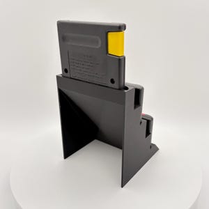 K&ouml;nnte beinhalten: Eine schwarze Videospielkassette steckt in einem schwarzen St&auml;nder. Die Kassette hat einen gelben Akzent. Der St&auml;nder hat ein Stufendesign und steht auf einer wei&szlig;en Oberfl&auml;che.