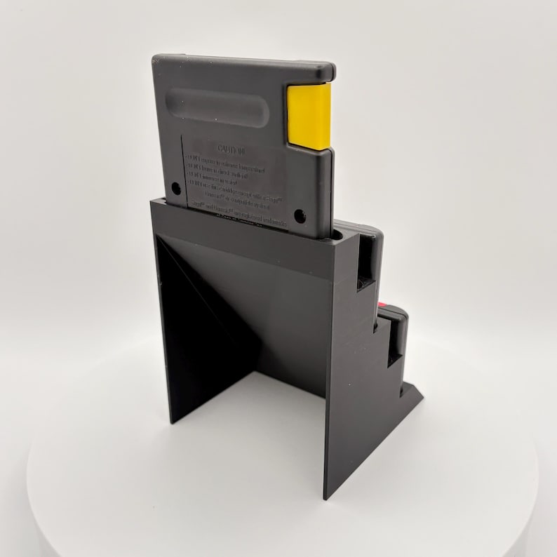 K&ouml;nnte beinhalten: Eine schwarze Videospielkassette steckt in einem schwarzen St&auml;nder. Die Kassette hat einen gelben Akzent. Der St&auml;nder hat ein Stufendesign und steht auf einer wei&szlig;en Oberfl&auml;che.