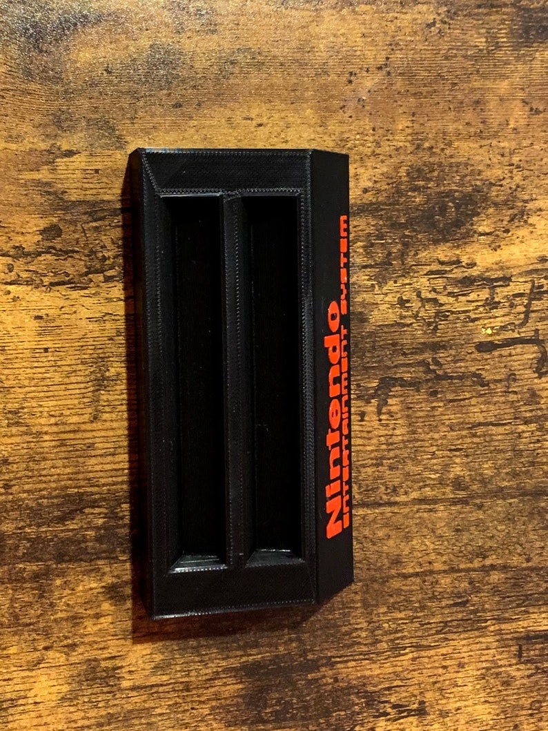 May include: Black plastic game cartridge holder with two slots and the text "Nintendo Entertainment System" in red.