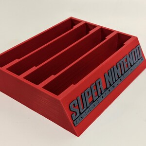 May include: Red plastic game cartridge holder with four slots and the text "SUPER NINTENDO" on the side.