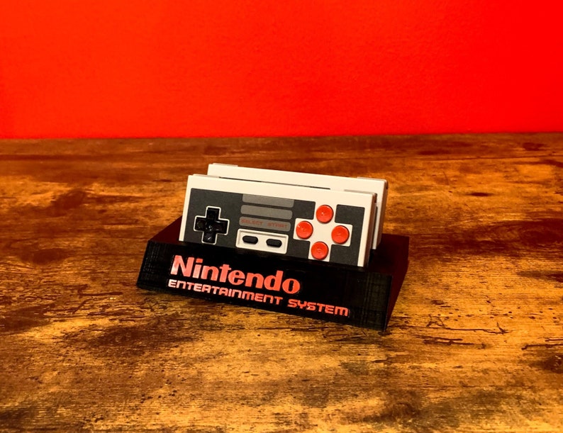 May include: A black stand with the text "Nintendo Entertainment System" on the front. Two gray and white Nintendo Entertainment System controllers are sitting on the stand.