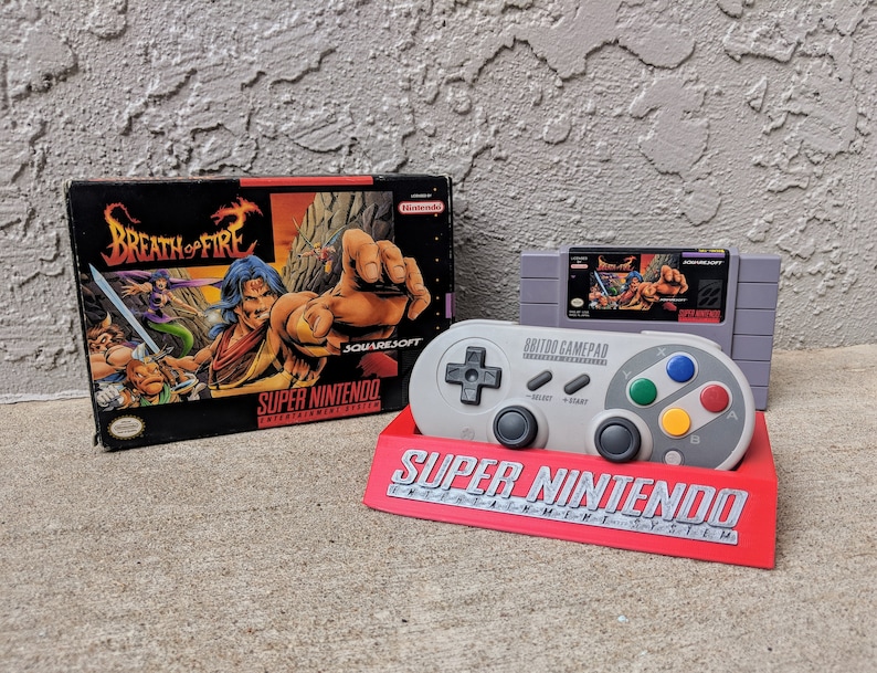 May include: A gray Super Nintendo Entertainment System controller with a gray cartridge for the game Breath of Fire II. The controller is sitting on a red stand with the words "Super Nintendo" printed on it.