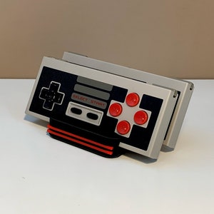 May include: Two gray and black Nintendo Entertainment System controllers with red buttons are stacked on a black and red stand. The controllers have the words "SELECT START" printed on them.